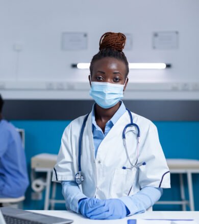Hospital medical specialist sitting at office table while wearing virus protection facemask and surgical gloves